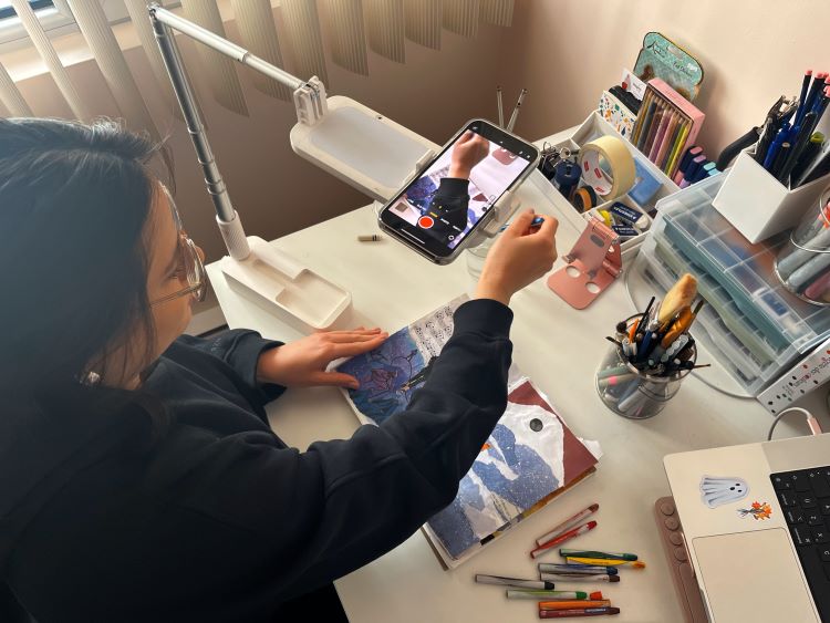 an artist working on a mixed medium collage project, her phone is positioned on a stand above the project filming the process.