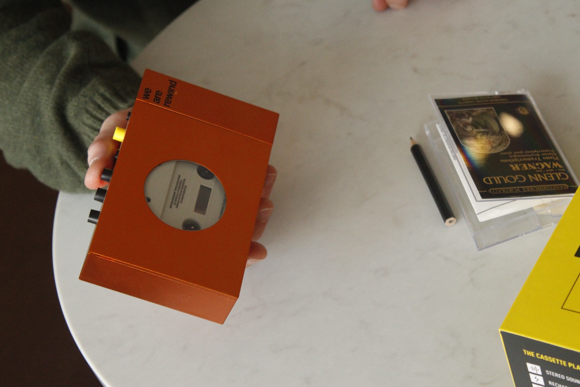 someone holding a new cassette tape player closed resting their arm on a table. a pencil and the cassette cover are also on the table