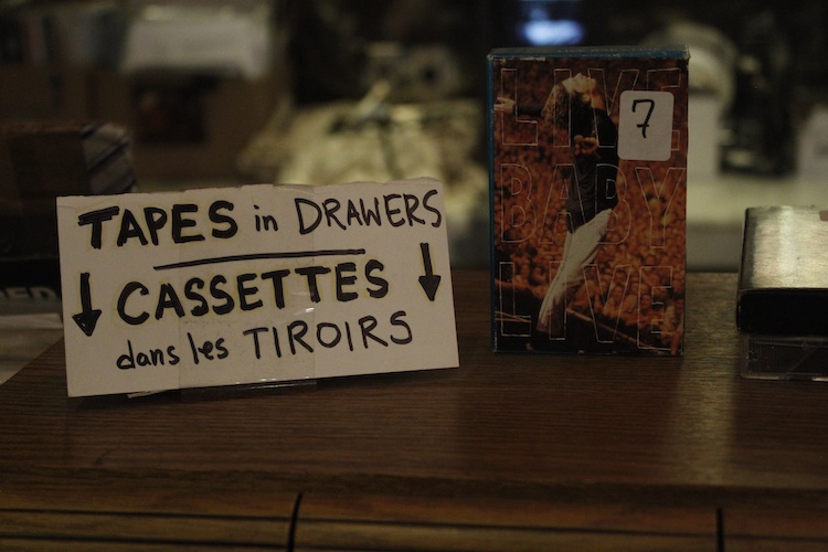close up of a sign that says "tapes in drawers. cassettes dans les tirroirs." next to the sign is a 7$ cassette.