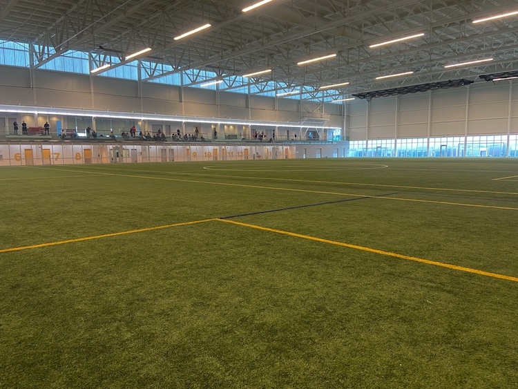A wide view of an empty soccer field with bright overhead lights and high ceilings. Spectators stand along an upper viewing area in the background.