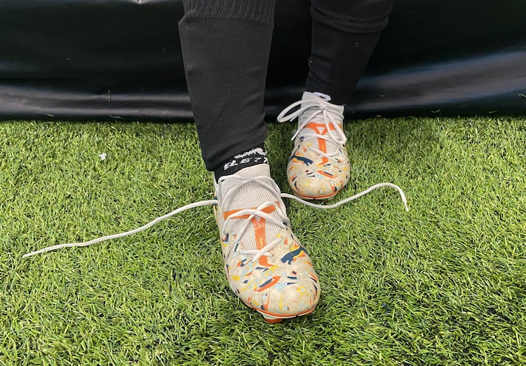 A close-up of a soccer player's feet on artificial turf. The player is wearing colourful patterned cleats with the laces of one untied.