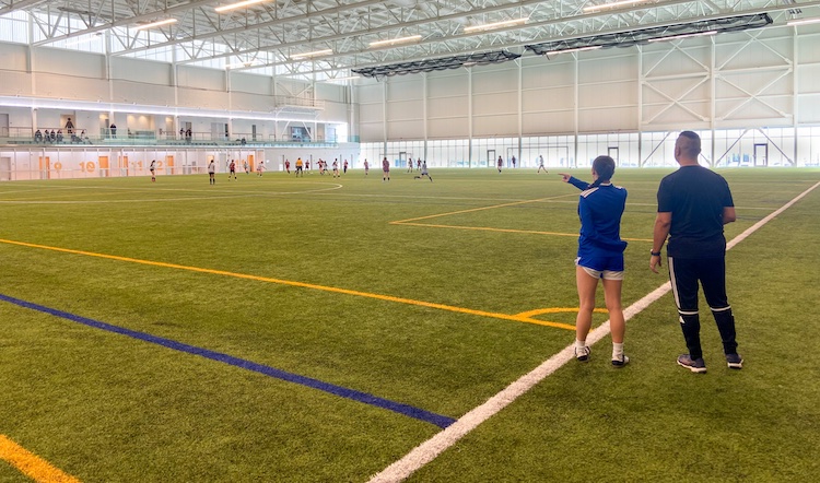 Two coaches are standing on the sideline of an indoor soccer game. The assistant coach points toward the field while the head coach watches, as a match unfolds in the background.