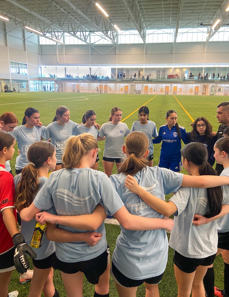 A group of young girls in soccer uniforms and their three coaches stand in a tight huddle with their arms around each other.